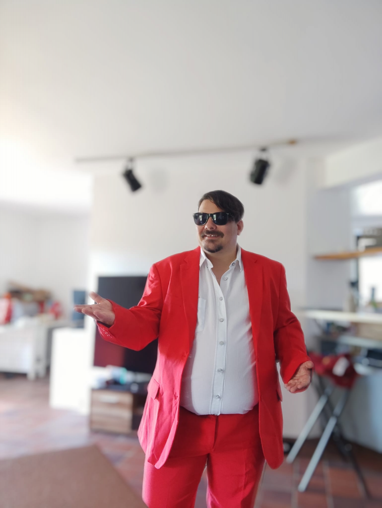 Auf dem Bild ist Timmo zu sehen. Timmo moderiert im Brauhaus u.a. das Pubquiz. Er trägt einen roten Anzug, ein weißes Hemd, Sonnenbrille und lächelt.